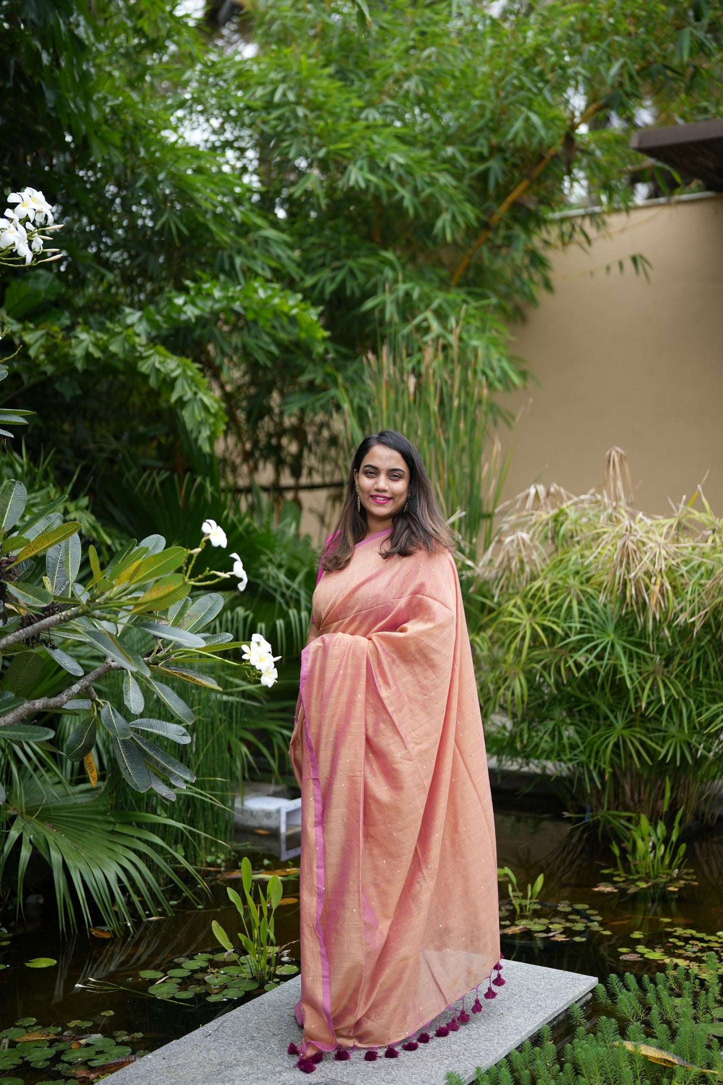 Woman in a peach saree standing in a garden with greenery and water features.