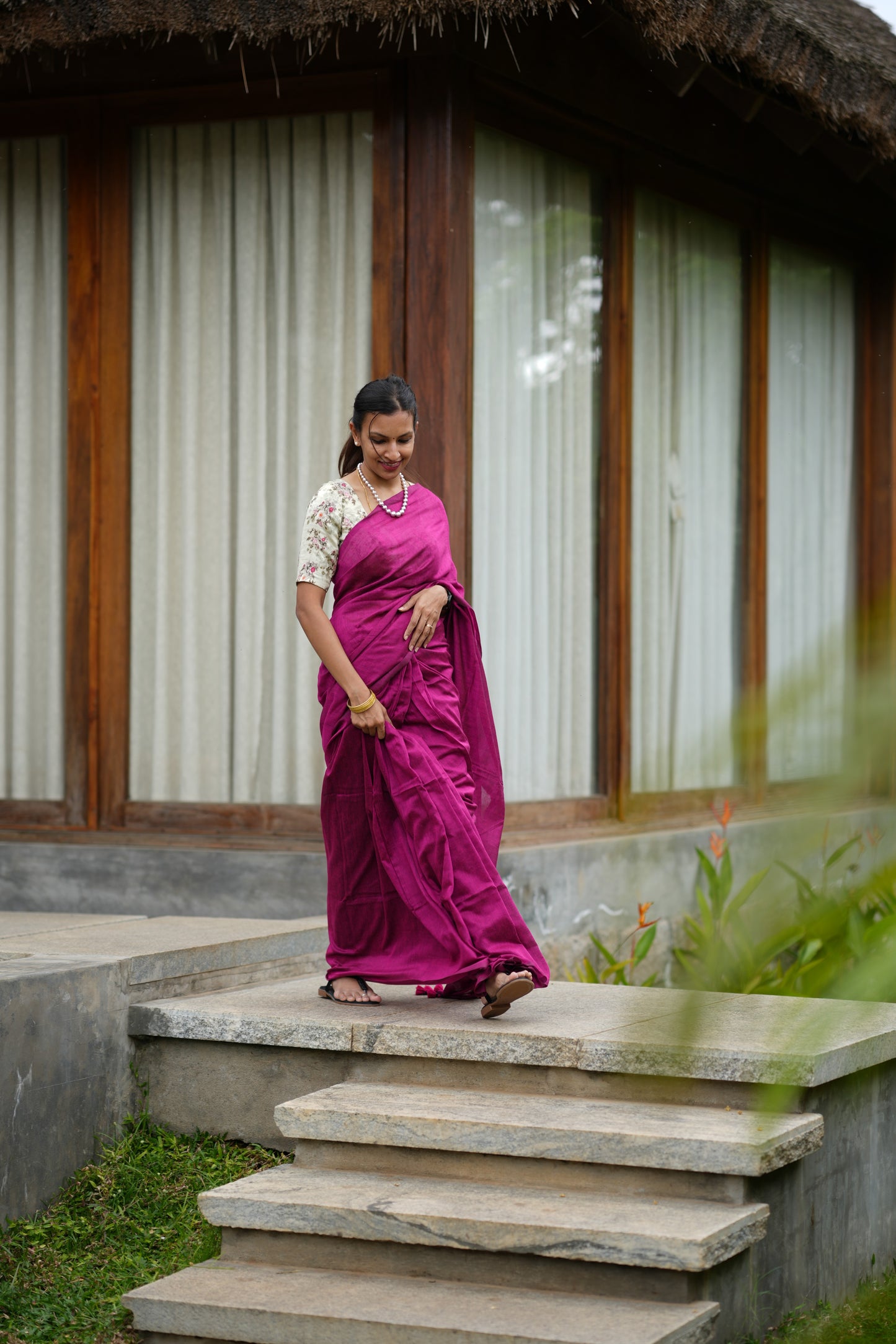 Woman in a pink saree walking up stone steps with a thatched-roof building in the background.