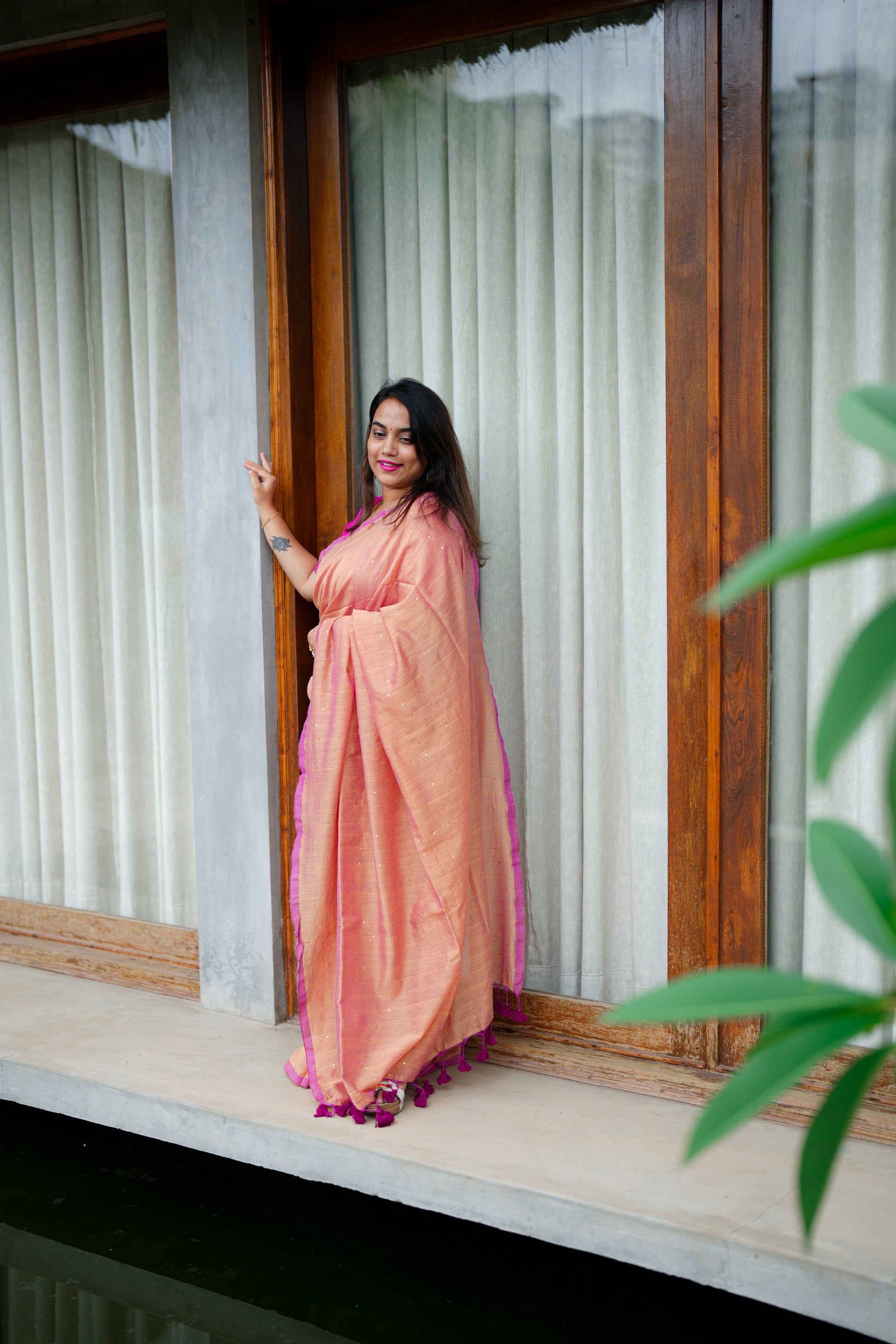 Woman in a peach saree standing by a window with curtains