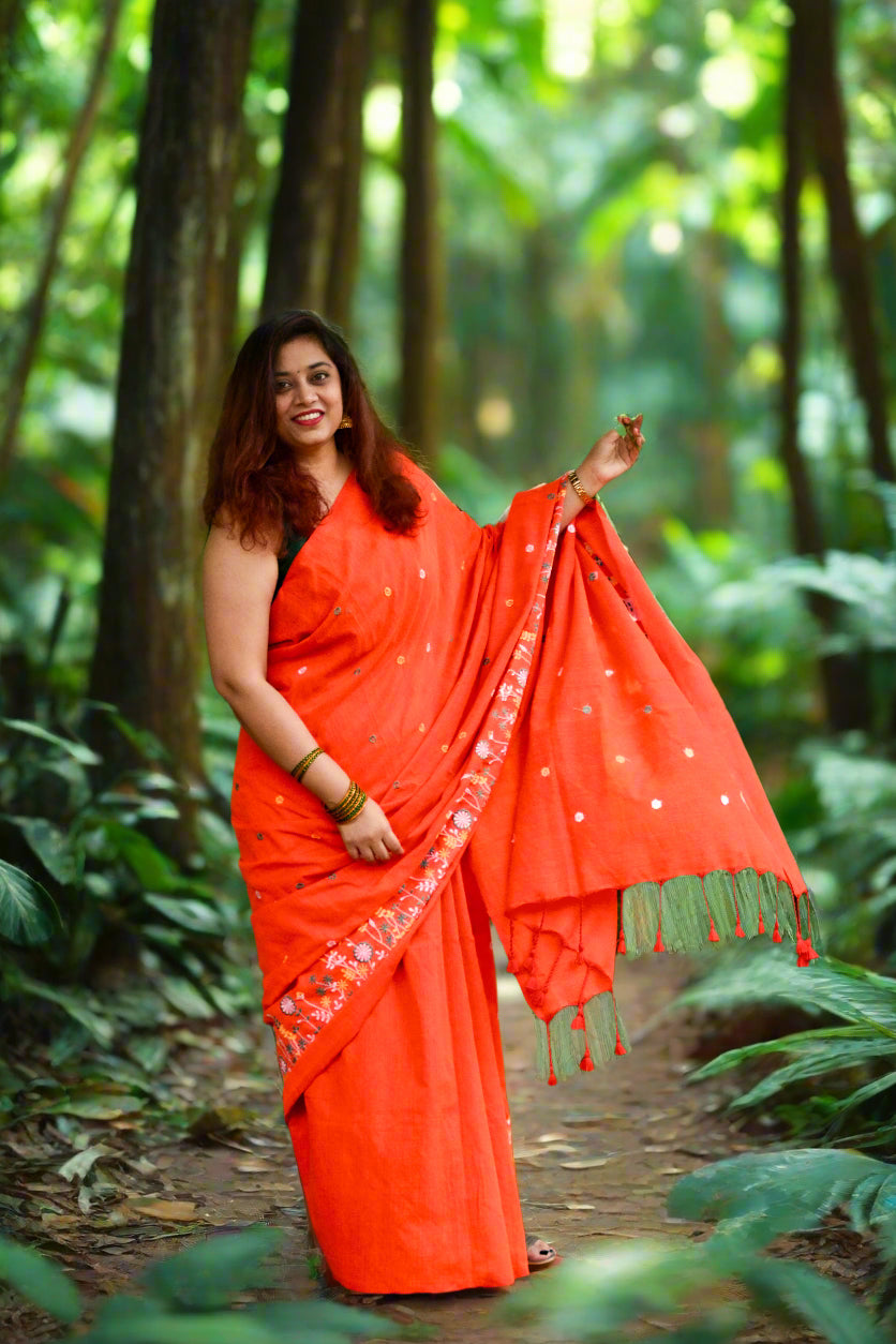 Woman in an orange saree standing outdoors with greenery in the background