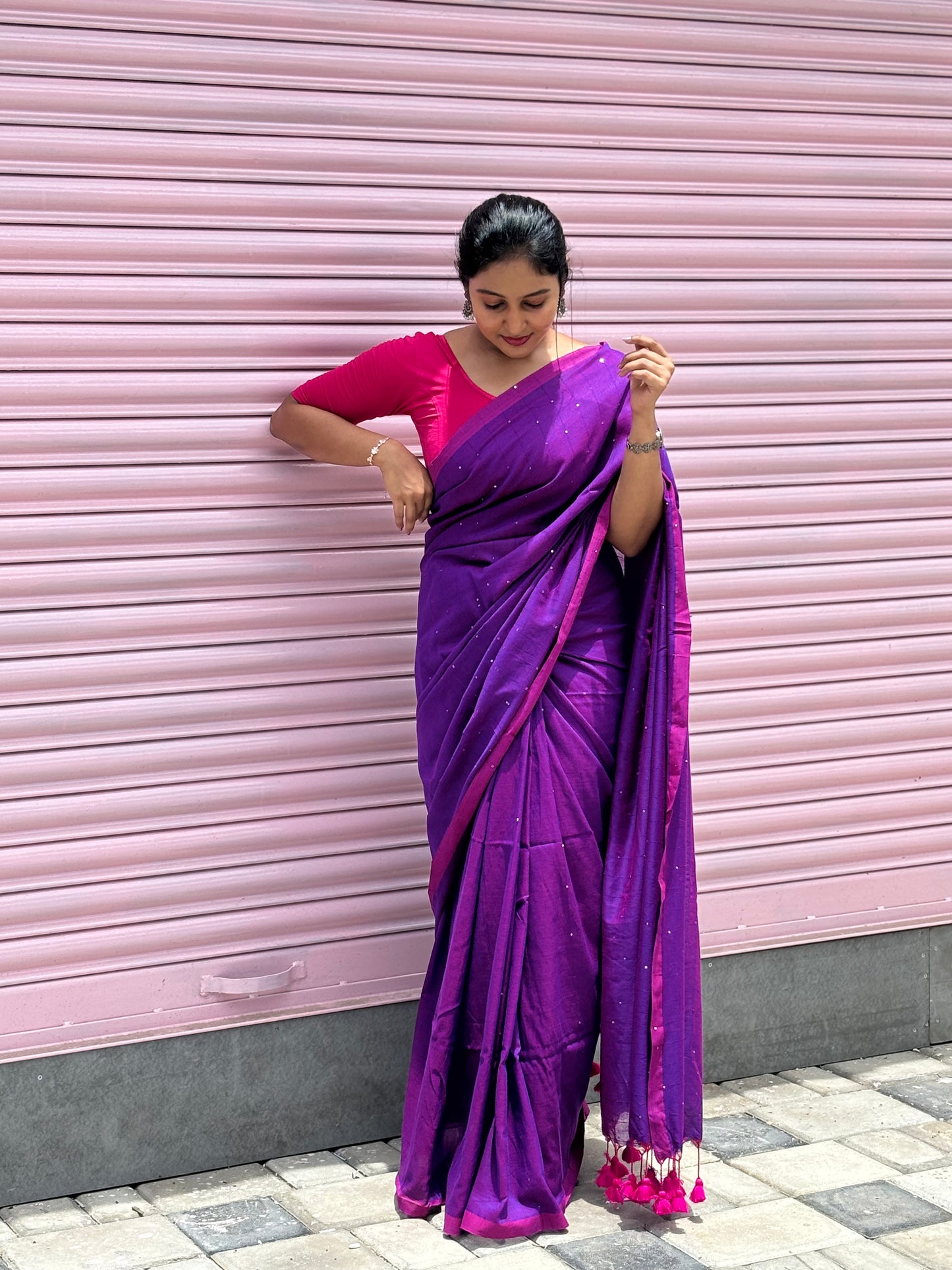 Woman wearing a purple saree with a pink blouse against a pink shutter background
