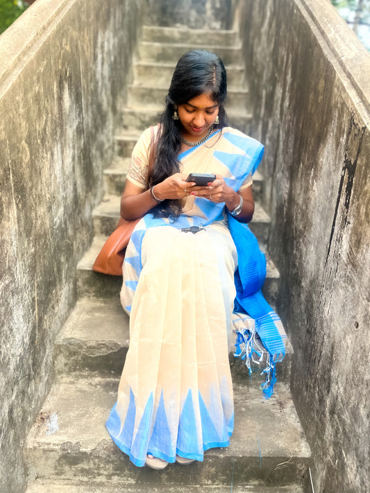 Woman in a blue and white saree sitting on steps using a phone.
