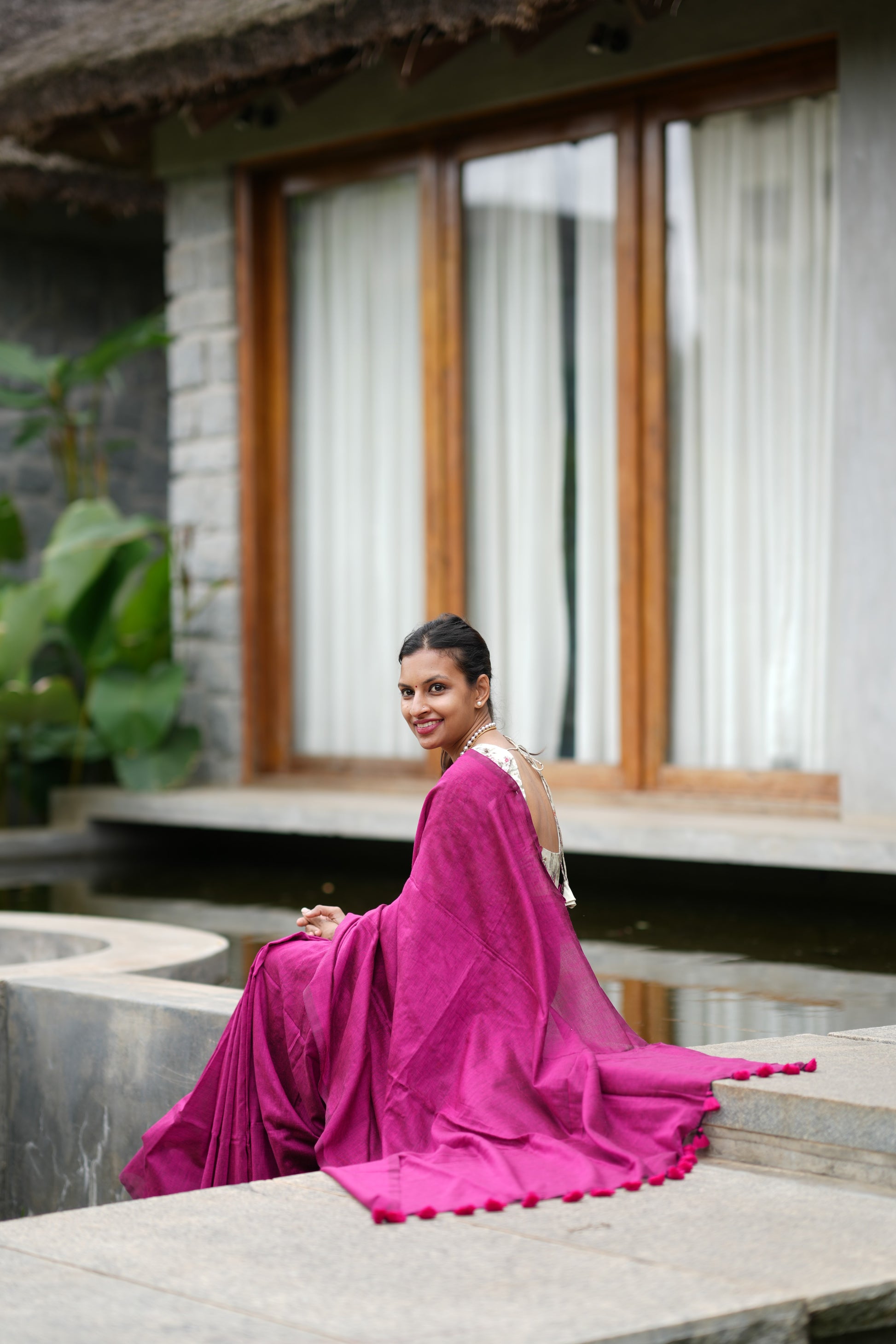 Woman in a pink dress sitting by a pool with a building and plants in the background