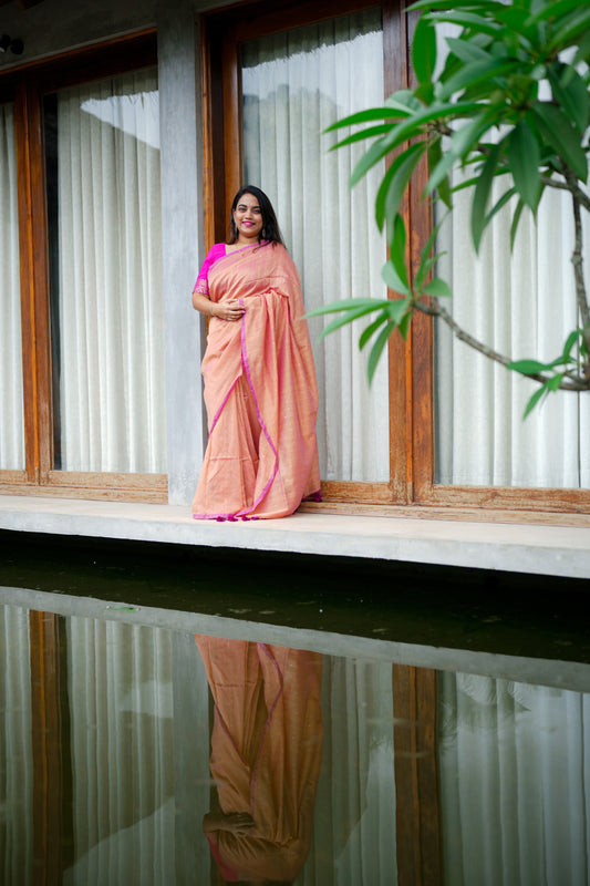 Woman in a peach saree standing by a window with a plant in the foreground